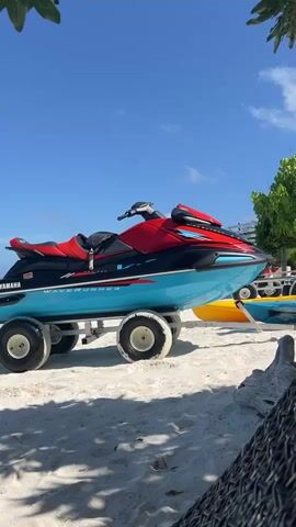 A blue and red Yamaha WaveRunner jet ski is on a trailer with large white wheels on a sandy beach. The jet ski is positioned to the left of the frame, facing right. The trailer is partially visible. Behind the jet ski, there is a glimpse of another watercraft, possibly a kayak, in yellow and blue. The ocean is visible in the background with clear blue water and a bright blue sky. There are some green leaves and branches in the upper left corner of the frame, suggesting foliage from a tree. In the lower right corner, a portion of a woven material, possibly a beach chair or hammock, is visible. The scene is brightly lit, indicating a sunny day.