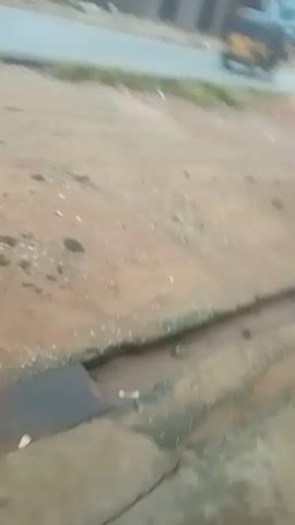 The video shows a roadside scene with a dirty, cracked concrete drain running along the edge of a dusty area. Several yellow auto rickshaws, also known as kekes, are driving on the asphalt road in the background. A person is briefly visible on the right side of the video, sitting or crouching near the edge of the road. The environment appears to be a typical urban or semi-urban street, with buildings visible in the background. The lighting suggests it is daytime, but the weather may be overcast.