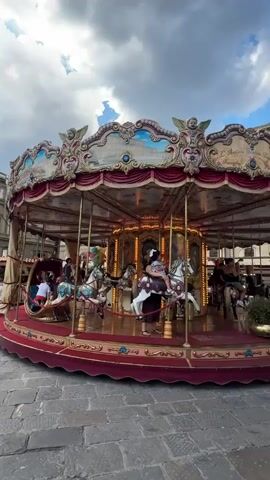 This is a video of a carousel with several people riding on it. The carousel is elaborately decorated with ornate gold trim and painted scenes of architecture and cherubs. It has red velvet drapes and a red and gold platform. The horses are colorful and detailed, some with elaborate tack. There is a crescent moon-shaped ride in the center.

The carousel is set up on a cobblestone street, suggesting an outdoor urban environment. In the background, there are buildings that appear to be historic architecture. The sky is partly cloudy, indicating it's likely daytime, possibly late afternoon or early evening, given the soft lighting.

The people on the carousel appear to be enjoying themselves. Some are children, while others are adults. One person in the crescent moon ride is looking out with a relaxed posture. The overall mood seems to be one of leisure and enjoyment.

While there is text visible on the carousel's canopy, it is too small and pixelated to read accurately. However, the style of the carousel and the architectural context strongly suggest a location in a historic European city, and the prompt specifically mentions Florence, Italy.