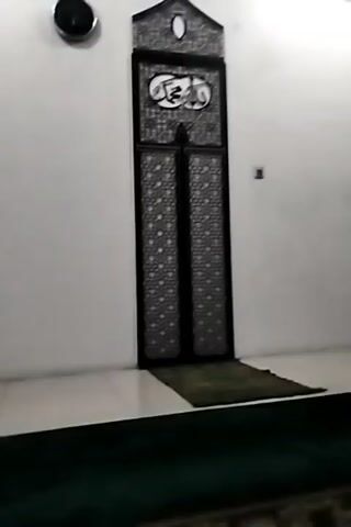 A video shows a close-up shot of a wall with a decorative door in the center. The door is tall and narrow, with intricate geometric patterns. Above the door, there is an Arabic inscription. To the left of the door, a small, circular object is mounted on the wall. A prayer mat is placed in front of the door. The lighting is dim, creating shadows on the wall. The video is short and appears to be a static shot.