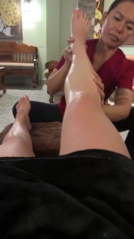 The video shows a person receiving a leg massage. The person getting the massage is lying on a bed covered with a brown towel and a black blanket. The massage therapist, a woman with dark hair wearing a red top and black pants, is sitting beside the bed and massaging the person's right leg with oil. The room has light green walls, a patterned tile floor, and wooden furniture in the background.