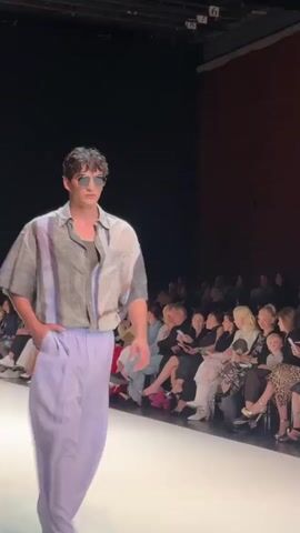 A male model walks down a runway wearing a light-colored, patterned short-sleeved button-down shirt over a darker tank top, and light purple wide-leg trousers. He has on sunglasses and his left hand is in his pocket. The runway is white. In the background, there is an audience seated on risers. The back wall of the venue is made of wood. The lighting is dim with spotlights on the runway.