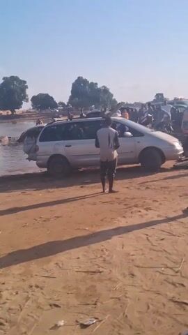 The video shows cars being transported across a body of water on a makeshift raft. The raft is made of wooden planks, and there are several people standing on it, guiding the cars and monitoring the process. The surrounding environment is a sandy or dusty area, with other vehicles and people visible in the distance. The sky is clear and blue, indicating a sunny day. The cars being transported include a silver station wagon, a red hatchback, and a dark-colored compact car. The people on the raft are dressed casually, and some are barefoot. The video appears to be taken in a developing country, given the simplicity of the transportation method and the landscape.
