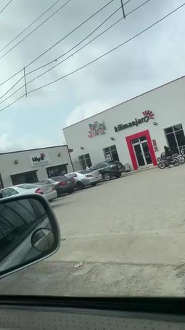 The video, filmed from inside a vehicle, shows a commercial building complex on a cloudy day. The main building on the right is white with red accents, displaying signs for "Pizza Jungle" and "Kilimanjaro." To its left is a smaller white building with a black roof, identified as "Kilimanjaro Grill." A paved parking lot fronts both buildings, where several cars are parked, including a silver sedan with its hood open and a black sedan. A white van with various stickers on its rear window, including "UNCLE YOKE'S HOT TOO," briefly passes in front of the camera, obscuring the view. Shortly after, a man wearing a t-shirt and shorts rides a red motorcycle across the scene, moving from left to right through the parking area. Power lines are visible against the sky. People are seen near the building entrances and in the parking lot.
