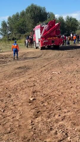 A group of people on horseback are gathered in a dirt area with some trees and a clear blue sky in the background. A red fire truck with a crane is parked in the middle distance. Several individuals in orange vests and blue shirts are present, some standing near the fire truck and others near the horses. One person in an orange vest and blue pants walks away from the camera towards the horses. An ambulance is partially visible on the right side of the frame. The ground is uneven and covered with dirt and some loose stones. The scene appears to be an outdoor event or possibly a training exercise.