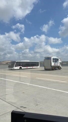 A time-lapse video shows an airport tarmac under a blue sky with scattered clouds. On the tarmac, there are several vehicles, including a white bus with red and black lettering, a white truck with a cargo container, and other ground support equipment. The camera appears to be positioned inside a building or vehicle, looking out a window, as evidenced by the reflections on the glass and the dark frame at the bottom of the screen. The video captures the movement of clouds and the general atmosphere of an active airport environment. No people are clearly visible or engaged in specific actions.