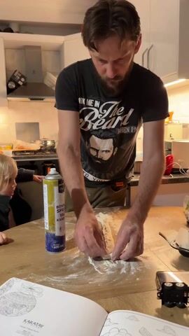 A man with a beard, wearing a black t-shirt, stands at a wooden table in a kitchen, working with a piece of dough covered in flour. He's focused on shaping it with his hands. Next to him on the table is a tall can of cooking spray. A child is partially visible to the left, watching the man. A cookbook titled "KARATAY" is open on the table, and a black object is near the edge of the table. The kitchen has white cabinets and a stainless steel range hood above the stove.