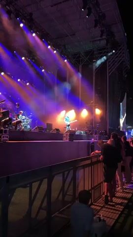 The video shows a live concert performance. A bald male musician wearing a dark blue t-shirt and jeans is playing a bass guitar on stage. He is standing on a platform in front of a wall of speakers. The stage is lit with purple and orange lights, and there is smoke or haze in the air. Several other musicians and their instruments are visible in the background, including drums. In the foreground, a metal barrier separates the audience from the stage. Several audience members are standing behind the barrier, watching the performance. One person is sitting down behind the barrier in the lower right corner of the frame. The overall atmosphere is that of a nighttime outdoor concert.