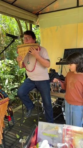 A man wearing glasses, a light brown t-shirt, and blue jeans sits on a stool and plays a pan flute. He has a necklace with a blue pendant. A young girl with dark hair and a brown long-sleeved shirt and blue jeans stands to his right, holding a small tambourine. She appears to be watching the man play. There is a microphone to the left of the man. Behind the girl is a table with a laptop computer. The scene appears to be outdoors, possibly under a canopy, with lush green foliage in the background. There are also several pan flutes and what appear to be CDs on display in the foreground.
