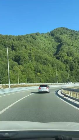 The video shows a view from inside a car traveling on a highway. The car is following a silver Volkswagen Golf. To the left of the highway, there is a green, tree-covered mountain. The sky is clear and blue. There are streetlights and guardrails along the highway. Other cars can be seen in the distance. The road appears to be curving to the right.