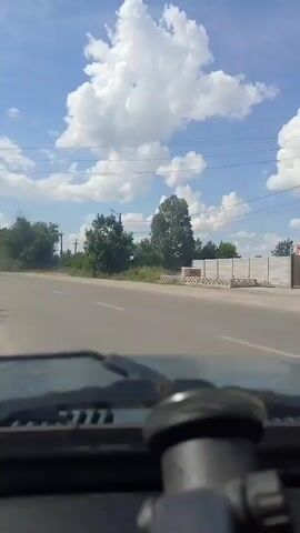 This is a first-person perspective video, likely captured from the dashboard of a car. The scene is a road on a bright, sunny day with scattered white clouds in a clear blue sky. The road appears to be a two-lane asphalt road, with white lane markings. On the right side of the road, there is a white fence with some decorative elements, and behind it, there are some buildings and trees. On the left side of the road, there are more trees and some power lines run overhead. The overall setting appears to be a rural or semi-urban area. The time of day is clearly daytime, indicated by the bright sunlight and the shadows. The weather appears to be fair and pleasant. There is no visible text in the video. The primary activity taking place is driving along this road. The focus of the foreground is the dashboard of the car, with a part of the steering wheel and a knob visible. The image quality is somewhat grainy, suggesting it might be from an older camera or phone.