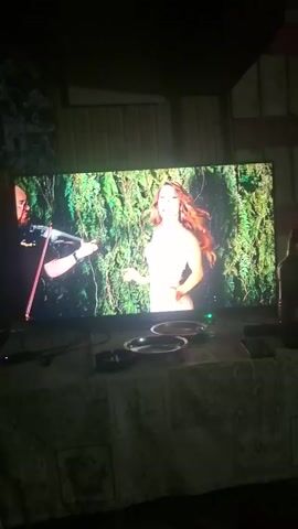 The video shows a TV screen displaying a music video. The video alternates between a man playing the violin and a woman posing. The woman poses in front of greenery, on a beach with blue water, and in other outdoor scenes. The TV is sitting on a table covered with a patterned tablecloth. On the table there are several ashtrays, a remote control, a phone, and a bottle of beer. The room is dimly lit.