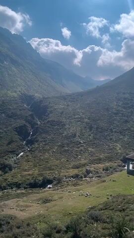 A vertical video pans across a vast mountain landscape under a partly cloudy blue sky. Lush green vegetation covers the slopes of the mountains, with a narrow stream winding through a valley on the left side of the frame. The sun is shining brightly, casting shadows on the mountains. The overall impression is one of a wild and undeveloped natural environment.