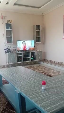 The scene presents a living room interior with the TV displaying a man in a red shirt and dark blazer. A large, gray table with a lower shelf sits in the foreground. A red and white object rests on the table. In the background, there is a light gray TV stand with various electronics on it, including the TV itself. Two small, white shelves hang on either side of the TV stand. The room is carpeted with a light brown rug, and the walls are painted in a neutral tone. The ceiling has a white, recessed design. A calendar hangs on the wall to the right of the frame.