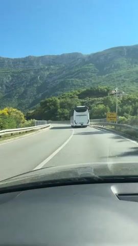 The video shows a view from inside a car driving on a paved road. In front, a large white bus is moving in the same direction. The road is straight and has a solid white line down the middle. Metal guardrails line both sides of the road. In the background, a large, green, mountainous landscape is visible under a clear, blue sky. A sign is positioned on the right side of the road that reads "Durmani 50 m". The camera is stationary, focusing on the road ahead. The car appears to be maintaining a consistent distance from the bus. Another car is seen briefly in the distance, in front of the bus.