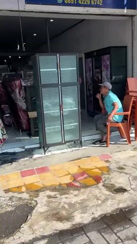 A man in a blue shirt and gray baseball cap sits on an orange plastic chair on a concrete sidewalk. To his left, a tall, glass-fronted cabinet with an aluminum frame leans precariously. To the left of the cabinet, two women are visible. One, wearing a white hijab and a patterned dress, stands and waves her right arm. The other, partially obscured, also appears to be wearing a hijab and a patterned garment. Behind the cabinet, an open doorway reveals the interior of a shop. A yellow spool is visible on a shelf inside the shop, and other items, possibly furniture, are also present. The ground outside the shop is a mix of concrete and brightly colored tiles. The overall scene suggests activity outside a store, possibly involving the movement or placement of the cabinet.