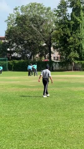 The video shows a cricket field with a player in grey walking towards the right, away from the camera. In the background, other players, some in teal shirts, are positioned near the wickets. The bowler is throwing the ball and the batsman is getting ready to hit it. A line of trees, some very tall, stands behind the players, with buildings visible through the trees in the distance. The grass is well-maintained, and the sky is partly cloudy. There's a practice net visible on the left side of the frame.