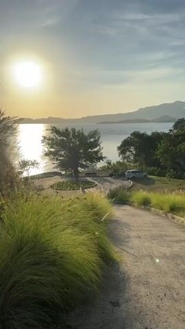 The video shows a landscape at sunset, focusing on a gently sloping paved path leading to a waterfront area. The bright sun shines over the sea, creating a shimmering effect on the water. In the foreground, clumps of tall, green grass line the path, with a few other trees and shrubs visible. Further down the path, there is a grassy area near the water where a tree provides shade. Several motorcycles and a silver car are parked there. The background features distant mountains, and the sky is a gradient of light blue and orange. The overall impression is one of peace and tranquility.