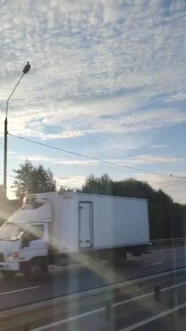 The video pans across a roadside scene under a sky filled with patchy white clouds. A highway runs horizontally through the middle of the frame, bordered by a metal guardrail. On the left, trees and vegetation line the roadside. A bright light source, possibly the sun, creates lens flares that streak diagonally across the image. In the distance, houses and trees are visible. A single utility pole stands on the right side of the road. Two small dark objects, possibly birds, are perched on a wire stretched across the frame.