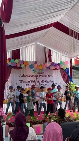 A group of young boys is performing a dance on a stage at an outdoor event. The stage is covered by a white and burgundy tent. There is a banner behind the performers that reads "Akhirussanah, Pentas Seni & Wisuda Tahfidz Al-Qur'an SDIT AZ-ZAHRA CURUG Tahun Ajaran 2022-2023." The boys are dressed in casual clothing, and some are wearing baseball caps. The audience, consisting of adults and children, is visible in the foreground. The overall atmosphere appears festive.