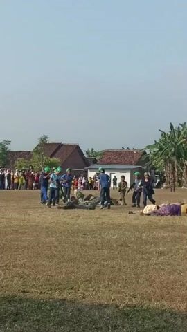 The video shows a group of people dressed in blue uniforms and green helmets on a dry, grassy field. Some of them are holding what appear to be toy rifles. Several people are lying on the ground, seemingly as if they have been knocked down. In the background, there are buildings with tiled roofs and trees. A crowd of onlookers is visible to the left of the scene. The sky is clear and bright, indicating a sunny day. The overall impression is of a staged event, possibly a reenactment or a performance.