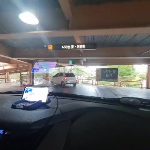 A dashcam view from inside a car shows a parking garage exit. The car is facing forward, with a GPS device mounted on the dashboard. A white car is visible ahead, exiting the garage. Signs in Korean and English indicate "EXIT." The environment appears to be an outdoor parking structure with concrete pillars and wooden beams. The time displayed on the car's dashboard is 4:52.