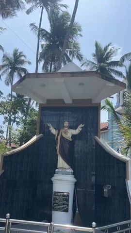 A video shows a statue of Jesus with outstretched arms, set against a backdrop of palm trees and a blue and white building with a cross on top, possibly a church. The statue is on a pedestal, with a black tiled wall behind it. The scene appears to be outdoors in a sunny location.
