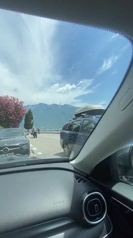 This is a first-person perspective video taken from inside a car. The first shot shows the view out the windshield, with a road, mountains, and a body of water visible. There are motorcycles on the road. The second shot is from the side window, showing the road and the body of water. The third and fourth shots are of two people inside the car. A woman in a blue bikini is in the foreground, and a man with curly hair is in the background. The woman is looking at the camera and making a peace sign. The car appears to be moving.