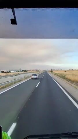 The video captures the view from the driver's seat of a truck. A GPS navigation device is visible on the dashboard, displaying a route. Outside the windshield, a highway stretches ahead with a white car in the distance. A side mirror reflects the highway and a golden sky. Another shot focuses on the side mirror, showing the road receding into the distance with a field and a body of water visible on the right. The final scene reveals a sunset with a bright sun illuminating the sky, casting a warm glow over the landscape. The overall impression is that of a peaceful drive through the countryside at dusk.