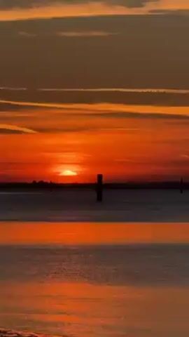 The video shows a sunset over water. The sun is a bright orange-red and is partially obscured by clouds on the horizon. The sky is a mix of orange, red, and gray, with long, thin clouds stretching across it. The water reflects the colors of the sky, creating a shimmering effect. There are several wooden posts in the water.