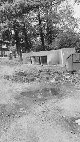 The video shows a dirt and grass area with several objects. In the background, there is a row of concrete structures that appear to be bins or dividers, with several large, square openings. Behind these structures, trees and foliage are visible. To the right of the concrete structures, a large metal dumpster is present, filled with debris. The foreground of the video shows a muddy patch with puddles of water and scattered debris. The overall scene appears to be an outdoor waste disposal or collection area, possibly in a somewhat neglected state, with evidence of water accumulation and general untidiness.