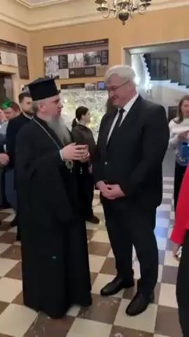 The video shows a brief scene in an indoor setting, possibly a lobby or hall, where three people are the main focus. A bearded man wearing a black religious robe and hat is speaking to a man in a black suit. A woman wearing a red jacket joins them. Several other people are visible in the background, including a child in a striped shirt. The walls have light beige tiling. There is a display panel with images on the wall to the left, and a staircase visible in the background leading upstairs. An ornate chandelier hangs from the ceiling. The main action involves conversation between the three individuals, with the man in the robe appearing to be the primary speaker.