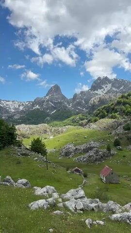 The video showcases a scenic landscape featuring two prominent mountains partially covered in snow, set against a blue sky adorned with scattered white clouds. In the foreground, there is a verdant valley with lush grass and patches of rocky terrain. A small stone building with a red roof stands alone in the valley. There's also some low level tree coverage in the valley. The camera pans upward and in, providing a wider view of the mountains before returning to its original position. The overall impression is one of untouched natural beauty.