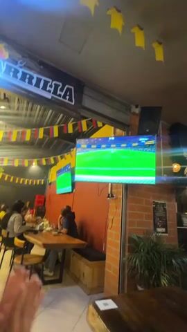 The video shows the interior of a sports bar. A television screen is visible in the upper left, displaying a soccer match. The room is decorated with flags from various countries, including Colombia. People are seated at tables, watching the game and socializing. The lighting is dim, with string lights adding to the atmosphere. The scene suggests a lively environment, possibly during a sporting event.