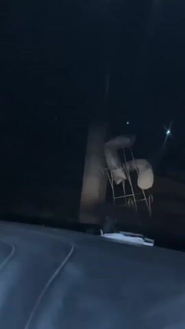 The video captures a dark, nighttime scene from the interior of a vehicle, likely a car, showing part of its dark dashboard in the foreground. In the midground, illuminated by an unseen light source, are several objects resembling bar stools or chairs, each individually wrapped in a light-colored, possibly white or beige, covering. These covered chairs are arranged on a metal framework. Behind them, a large, light-colored concrete pillar or similar structure is visible. Further in the background, several small, bright points of light are scattered against the dark sky, possibly distant streetlights or other light sources. No specific activity is occurring, and there are no people visible. The video was recorded in Idi Rayeuk, Indonesia.
