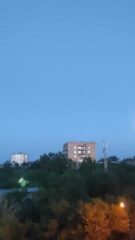 The video shows a twilight scene. The sky is a deep blue with a hint of lighter blue near the horizon. Two apartment buildings are visible amongst trees. The building on the left is taller and has several lit windows. The building on the right is shorter and also has lit windows. A telecommunications tower is visible behind the taller building. A street light with a visible bulb is at the top right of the frame. The trees are dense and dark green, with some autumnal-colored foliage on the right. The scene appears to be from a slightly elevated perspective.