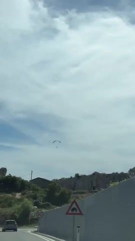 A paraglider is seen in the distance against a cloudy sky. In the foreground, there is a road with a speed limit sign and a tunnel warning sign. A white concrete wall is visible on the right side of the road. Trees and a building are situated on a hill in the background.