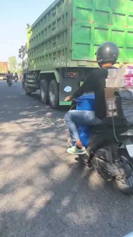 The video shows a busy street scene, likely in Southeast Asia, with heavy traffic. In the foreground, a black motorcycle, possibly with a rider wearing dark clothing and a helmet, is moving forward. To the left, another motorcycle, also with a rider, is positioned slightly ahead and to the left. Further ahead, a large green dump truck dominates the scene, traveling in the same direction. To the right of the dump truck, a black pickup truck is visible, also in motion. The road surface appears to be asphalt, with visible road markings. The environment suggests an urban or semi-urban setting with trees and some buildings in the background, under a clear blue sky. The overall impression is one of moderate to heavy traffic congestion.