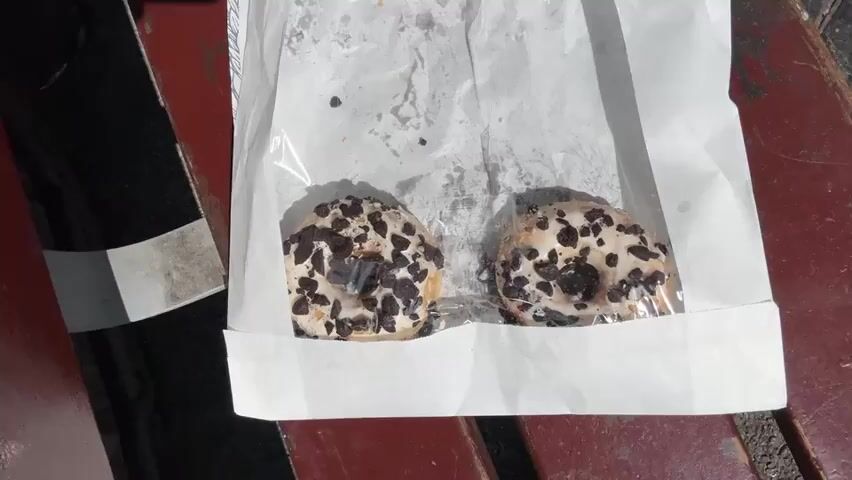 A person wearing sunglasses and a light blue shirt is shown. They are holding a donut covered in white icing and dark chocolate chunks, with a bite taken out of it. The donut is inside a plastic bag. The person is outdoors, with a blue sky and trees visible in the background. A market sign with red lettering is also visible. The video seems to be shot from a low angle, looking up at the person and the donut.