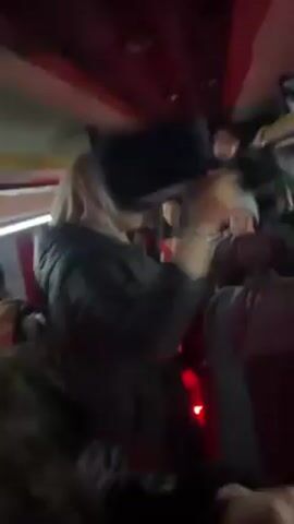 The video shows a crowded bus with people dancing and cheering. The interior of the bus has red and white accents, with bright lights along the ceiling. The passengers are wearing jackets and hats. One person is holding a black bag above their head. A bright red light is visible at the bottom of the frame. There appears to be loud music playing.