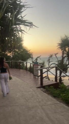 The video shows a beach scene during sunset. The camera appears to be moving along a paved path. There are green leafy trees on either side, partially obscuring the view of the ocean.  A wooden boardwalk with metal railings is visible, leading towards the beach.  The beach has rocks and waves.  Two girls, one with long dark hair wearing a white top and skirt and the other with blonde hair wearing a green top and blue pants, walk from behind a large rock towards the beach.  The sky is a mix of orange and grey.
