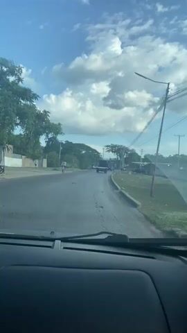 The video shows a view from inside a vehicle looking out through the windshield. It is daytime with a partly cloudy sky. The vehicle is on a paved road, which appears to be a highway or a main road. There are trees and vegetation along the sides of the road, as well as buildings and some utility poles with wires. There are other vehicles on the road, including a truck in front of the vehicle and a bus further down the road. A person on a bicycle is also visible on the left side of the road. The road appears to be in a somewhat rural or suburban area. The video is a continuous shot, with no apparent cuts or edits.