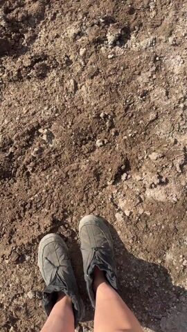 The video begins with a close-up shot looking down at the feet of an individual, wearing dark, sturdy boots, standing on heavily muddied ground. The mud is a rich brown color, mixed with dirt clumps and small stones, and appears uneven and soft.

As the camera slowly pans upward and backward, a wider scene is revealed. The path ahead is a deeply rutted, unpaved road, entirely covered in thick, wet mud. Numerous deep tire tracks crisscross the path, many of them filled with murky water, forming long, narrow puddles. Along both sides of the muddy path, substantial banks of dirty snow are visible, suggesting either late winter or early spring with melting snow.

In the distant background, several houses or buildings with peaked roofs can be seen under a clear, bright sky. To the right side of the path, a light-colored vehicle, possibly a delivery van or truck, is partially visible. Text is discernible on the side of the vehicle, including the Russian words "ОСТРОВ" (meaning "island") and "ТИПОГРАФИЯ" (meaning "printing house"). The sunlight is strong, indicating it is daytime, and reflecting off the wet surfaces of the mud and water. The camera continues to pan slightly, emphasizing the challenging, muddy terrain before slowly panning back down to a closer view of the extensive mud and tire tracks.