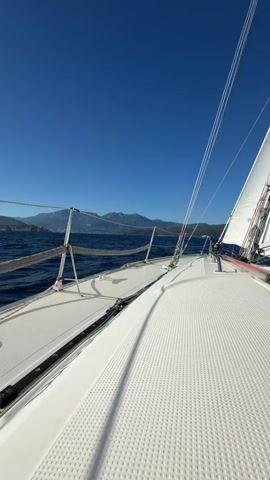 The video shows a first-person perspective from a sailboat moving on the ocean. The view is of the starboard side of the boat, looking forward and slightly to the left. The deck of the sailboat is white and textured, with a railing running along the side. In the distance, there is a coastline with mountains under a clear blue sky. The water is a deep blue and appears somewhat choppy. The sail of the boat is visible on the right side of the frame, white with red accents. The camera is looking out towards the horizon.