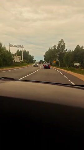 The video captures a view from inside a car driving on a highway. The dashboard and radio display are visible in the foreground. The radio is playing music, with song titles like "Cyberpunk 2077" displayed. The road ahead shows several cars and a truck. On the left side of the road, there is a large sign with white lettering on a white structure, and trees line both sides of the highway. The sky is overcast.