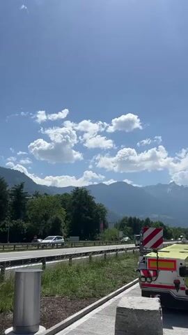The video shows a highway with cars driving along it, with mountains in the background. The sky is blue with white clouds. There is a rest stop called "Rheintal" to the right of the highway. There is also a guardrail, some grass and a trash can visible in the foreground.