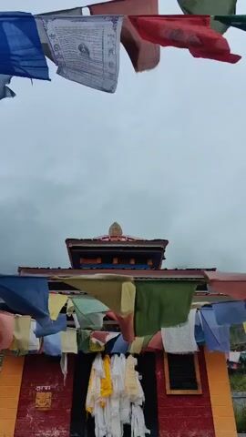 The video shows the front of a small building, possibly a temple or shrine. Prayer flags in various colors, including green, yellow, blue, and red, are strung across the front of the building and flutter in the wind. These flags appear to be made of cloth and have religious symbols or writings on them. In front of a dark doorway, several white and yellow cloth items are hanging, possibly offerings or prayer scarves. The building itself has a red facade, with some yellow paint visible on the sides. The roof is dark with some decorative elements. The sky is overcast and appears to be cloudy or foggy. The overall scene suggests a remote or mountainous location, consistent with a place where prayer flags are commonly used.