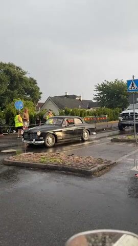 A black vintage Volvo is driving on a wet road. Several people are standing on the side of the road, some of whom are wearing yellow vests. A bicycle is leaning against a fence. There are houses and trees in the background. The sky is overcast.