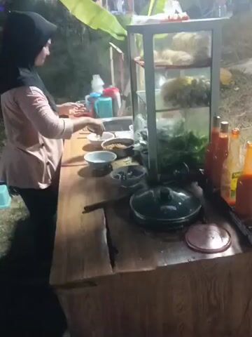 The video shows a woman wearing a hijab and a light pink top, black pants, and black shoes standing behind a food stall. She is leaning over the counter and appears to be preparing food. The stall has a wooden counter with various items on it, including bowls, bottles of condiments, and a covered pot. Behind the counter, a glass display case shows ingredients such as noodles, meatballs, and leafy green vegetables. In the background, there are banana leaves, a tent, and other people, suggesting an outdoor event or market. The lighting suggests it is evening or nighttime.