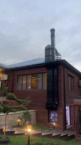 The video shows a wooden building with a large black chimney stack on the side. The building has several windows, some of which are lit from within. There are outdoor lights on wooden steps leading up to the building. A small tree is planted to the left of the steps. The sky is cloudy.
